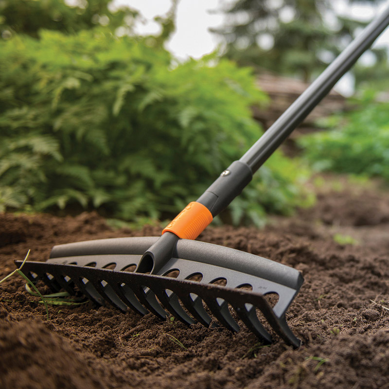 Musta harava oranssiliitoksella mullalla puutarhapenkissä, vihreitä lehtiä taustalla.
