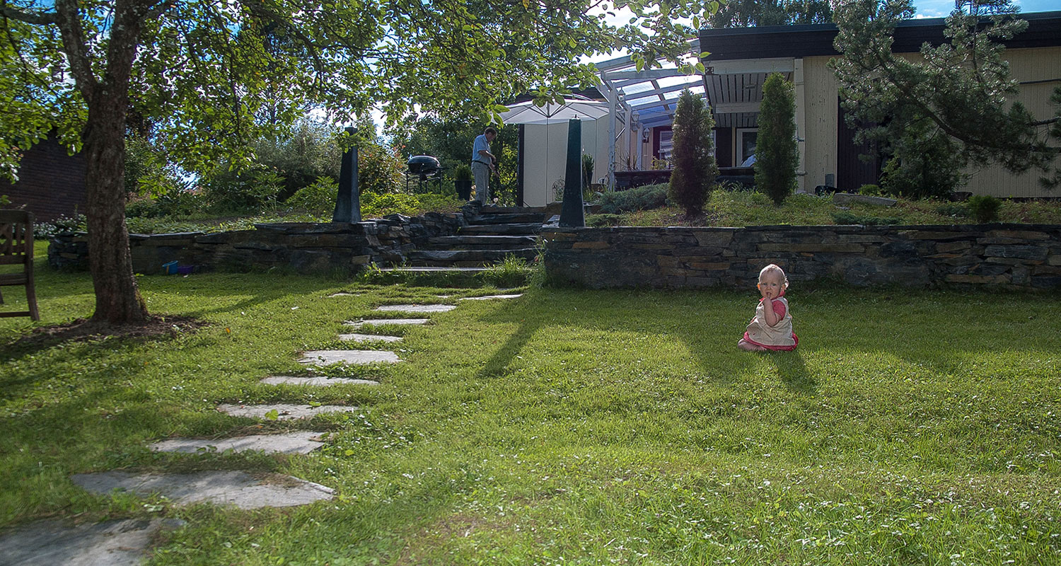 Vihreä piha kivipolkuineen, lapsi istuu nurmikolla, mies grillaa terassilla.