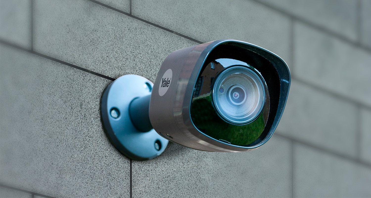 Sinertävä Yale-valvontakamera harmaalla laattaseinällä, linssi heijastaa vihreää nurmikkoa.