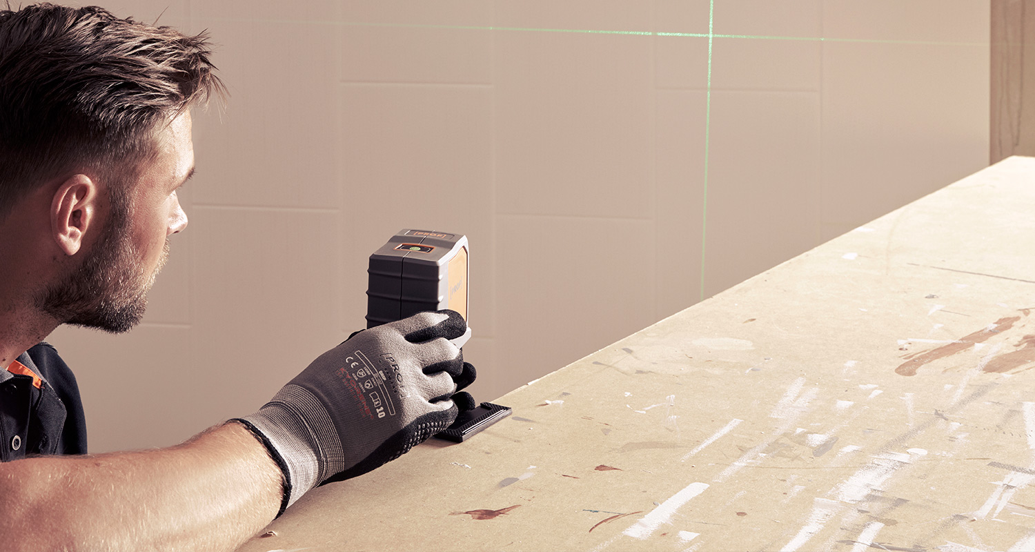 Harmaa-oranssi laserlinjalaite vihreällä ristivalolla työtasolla, suojakäsineinen käsi säätää