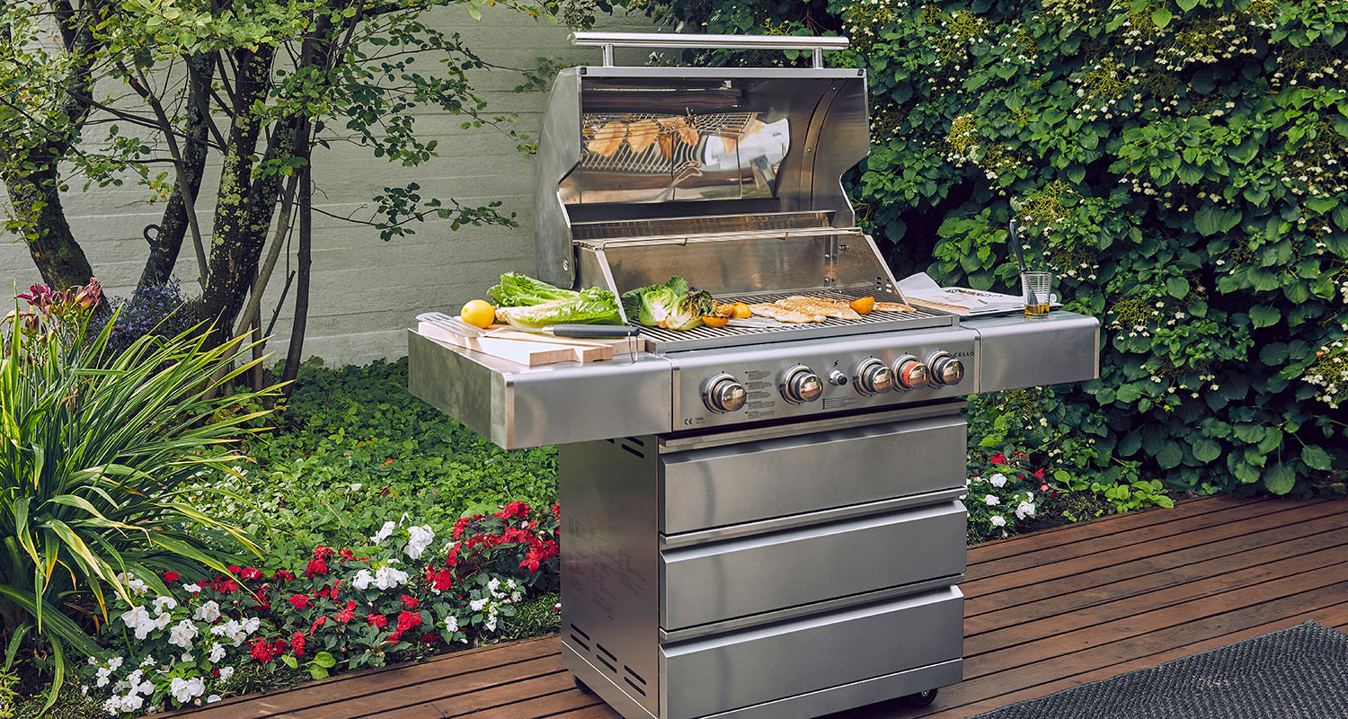 Ruostumateräksinen kaasugrilli puisella terassilla, ympärillä vehreää puutarhaa, punavalkoisia kukkia