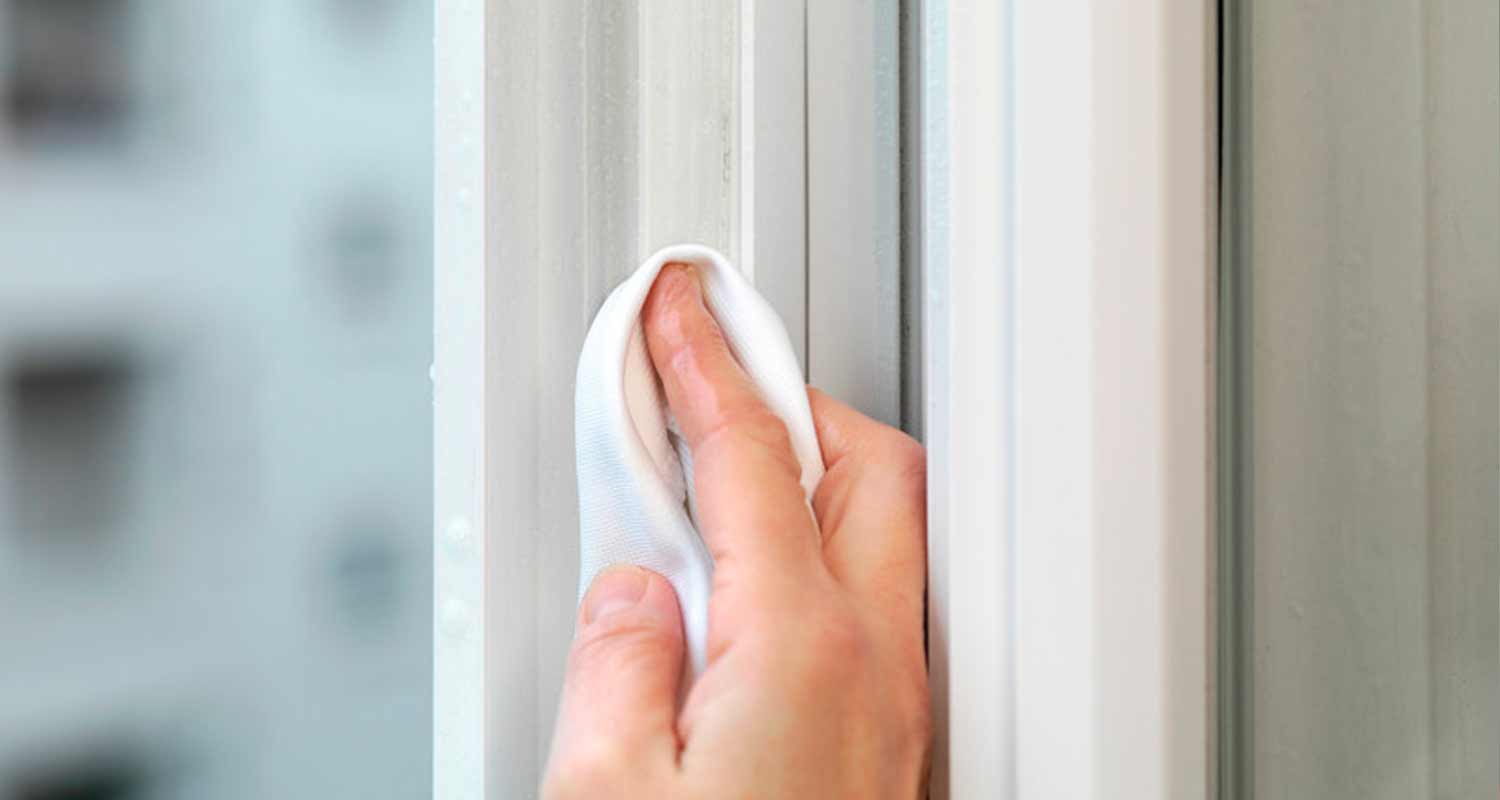 Käsi pyyhkii valkoista ikkunankarmia puhtaalla mikrokuituliinalla.