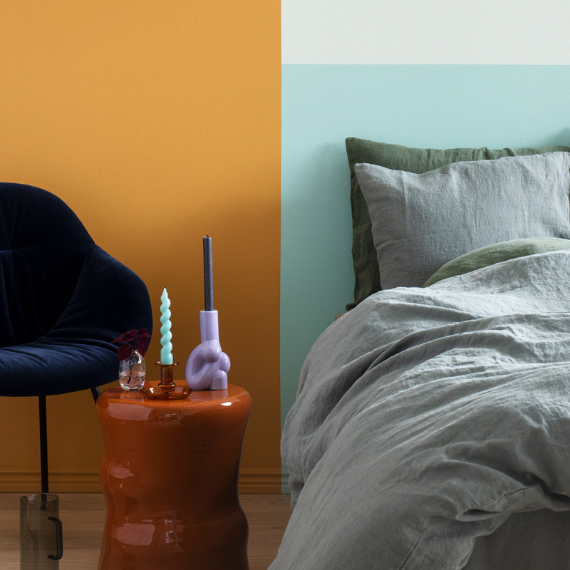 Kuvassa maalattu seinä pirteän keltaiseksi. Kuvasta löytyy myös musta jakkara ja pieni pöytä joka on sängyn vieressä. 