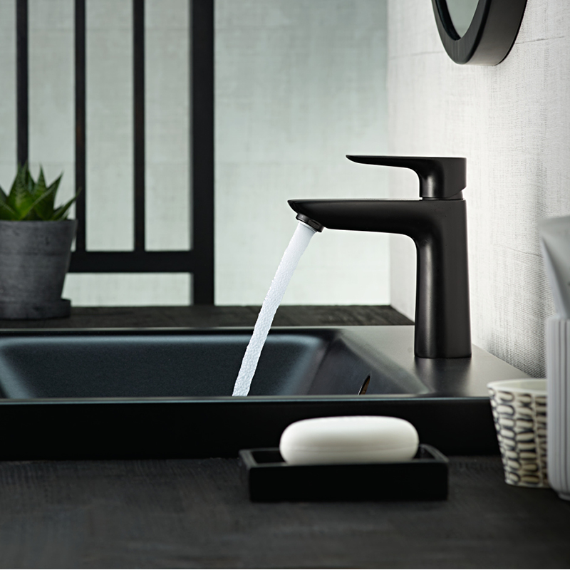 Kylpyhuoneessa musta hana valuttaa vettä mustaan altaaseen, edessä valkoinen saippua ja mehikasvi