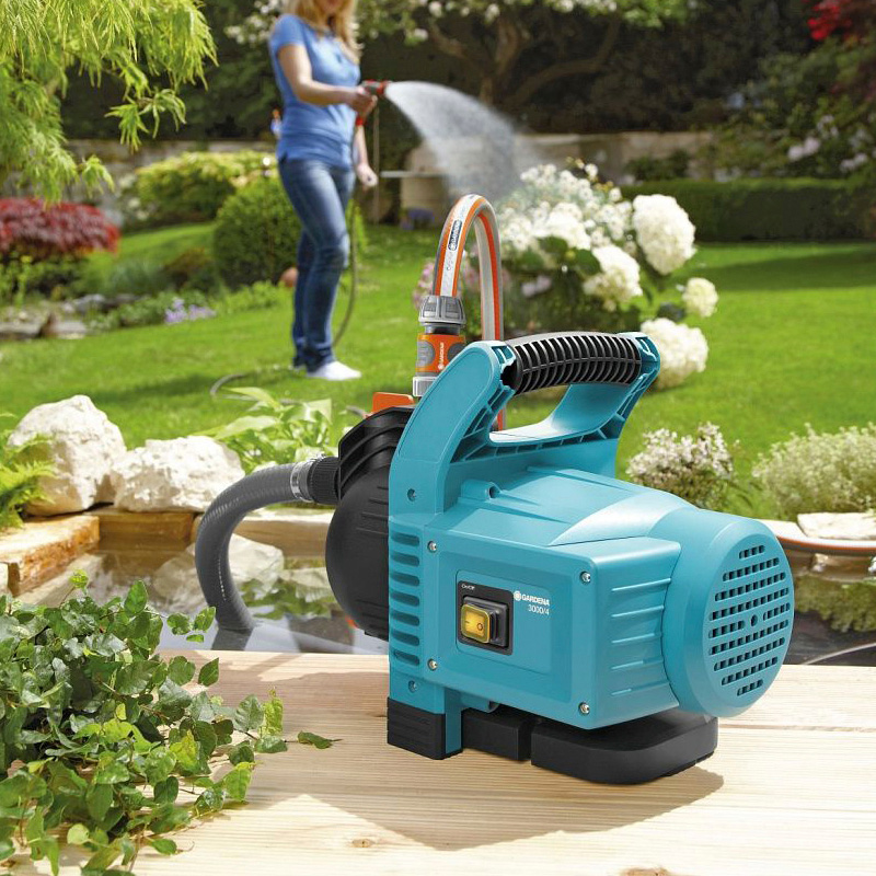 Turkoosi puutarhapumppu puupöydällä, taustalla nainen kastelee vihreää puutarhaa.