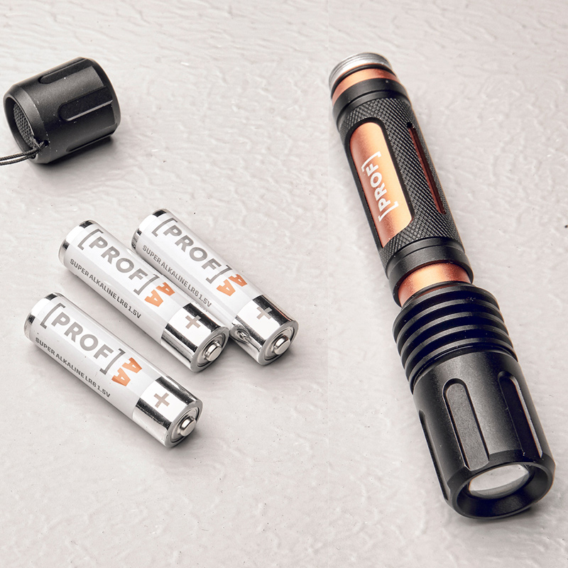 Musta-oranssi metallinen taskulamppu ja kolme valkoista PROF AA-paristoa vaalealla pinnalla