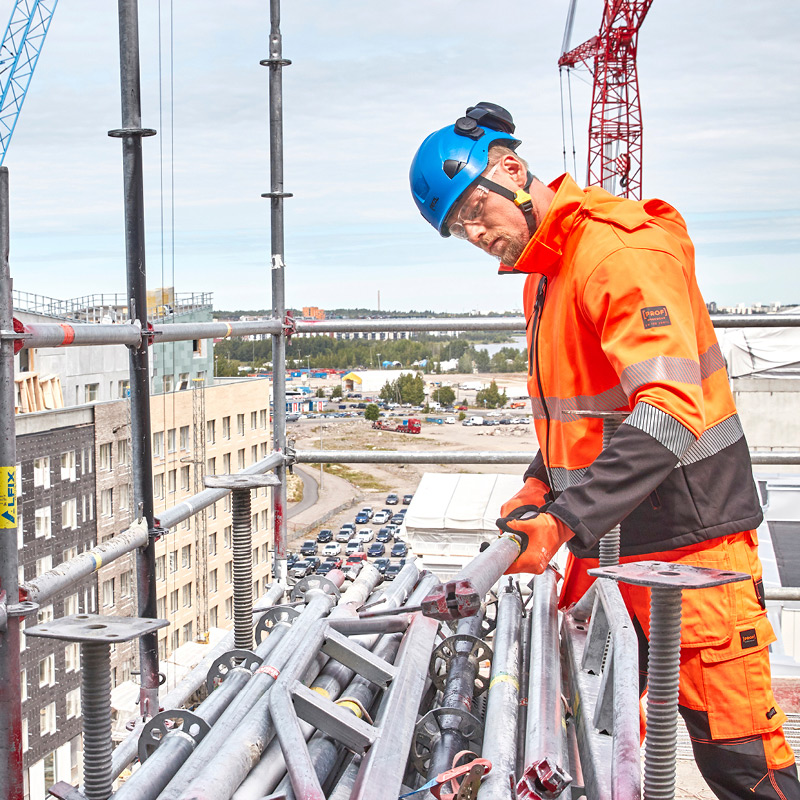 Rakennustyöläinen oranssissa haalarissa ja sinisessä kypärässä nostaa telineputkia työmaalla.