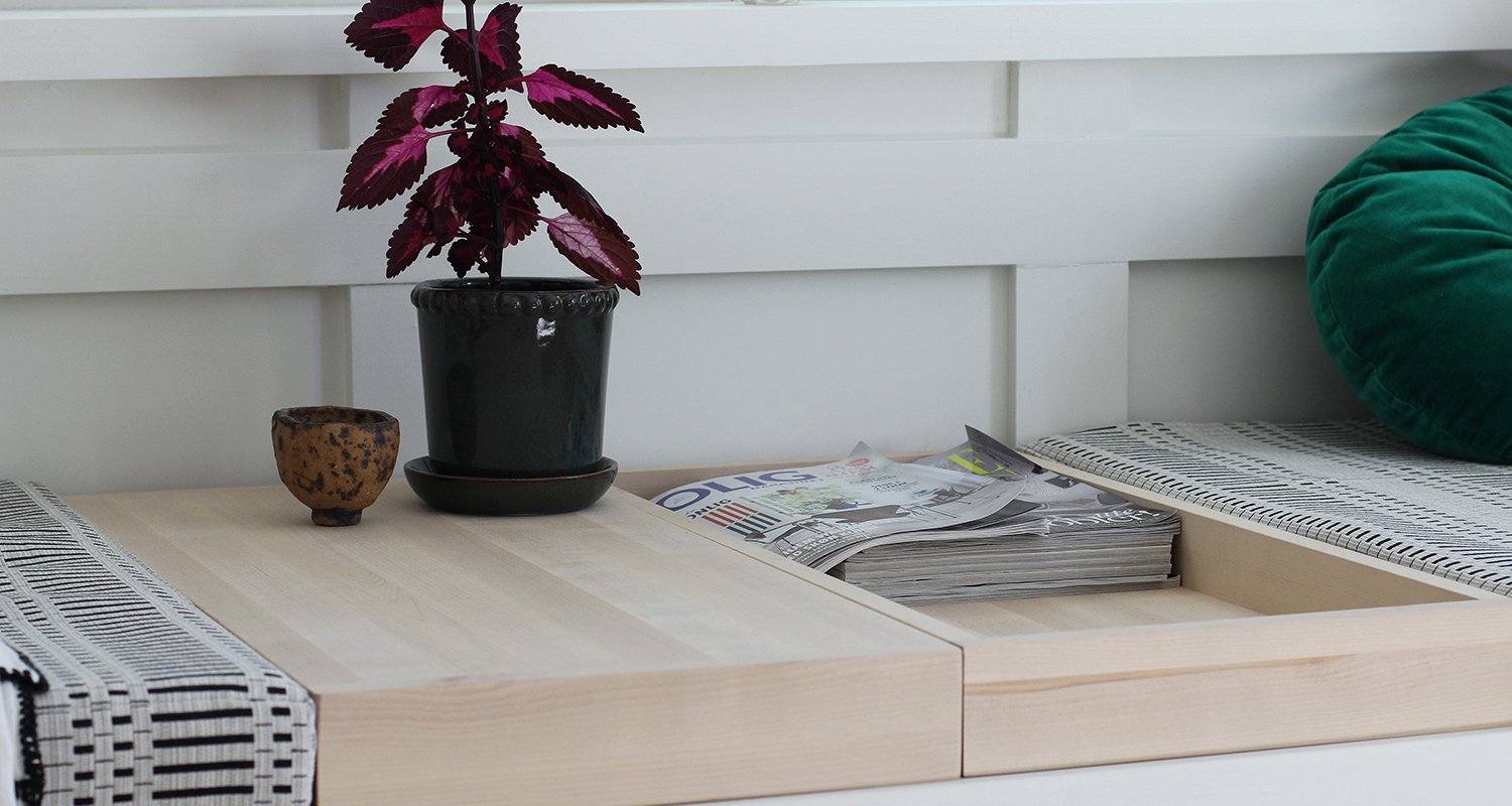 Vaalea puupöytä avattavalla kannella, sisällä pino lehtiä. Päällä violetti huonekasvi tummassa ruukussa.