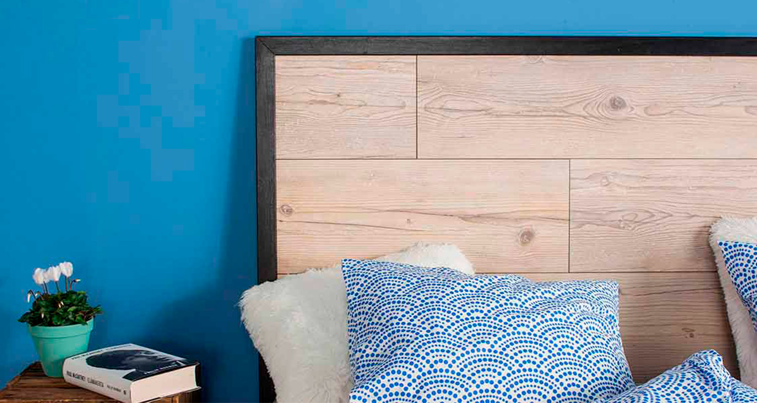 Sininen seinä, puinen sängynpääty, sinivalkoinen tyyny ja vihreä kukkaruukku yöpöydällä.