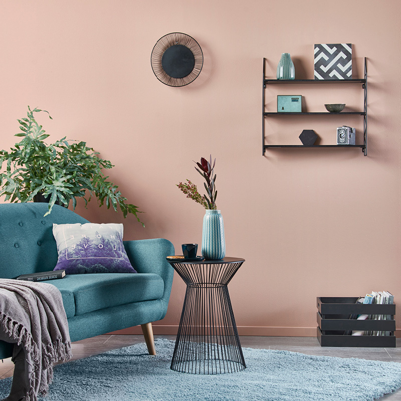 Vaaleanpunainen seinä, turkoosi sohva, musta rautapöytä sinivihreällä maljakolla ja hylly koristeineen.