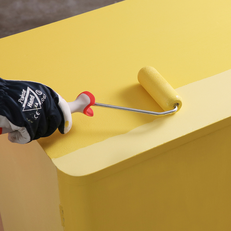 Kuvassa maalataan lipastoa keltaiseksi kalustemaalilla. Maalaajalla kädessä pieni tela ja työkäsine.