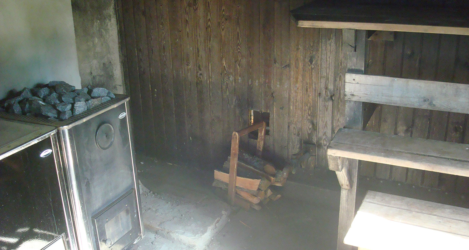 Saunan musta metallikiuas harmailla kivillä, puukori polttopuilla ja puulauteet puuseinän edessä.