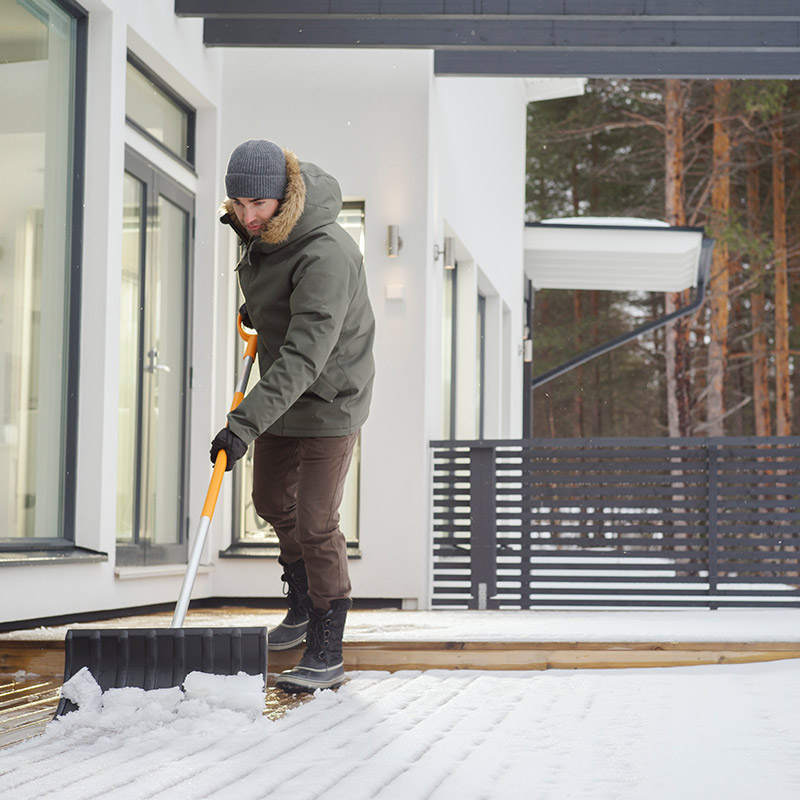 Henkilö lapioi lunta modernin valkoisen talon edustalla oranssinkahvaisella lumikauhalla