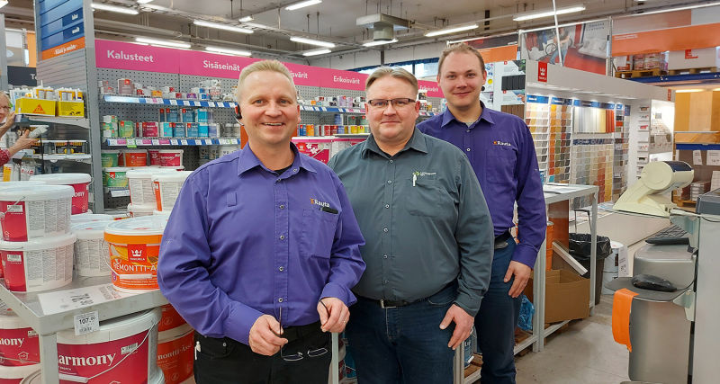 Kolme rautakaupan myyjää violeteissa paidoissa maali- ja väripurkkihyllyjen edessä.