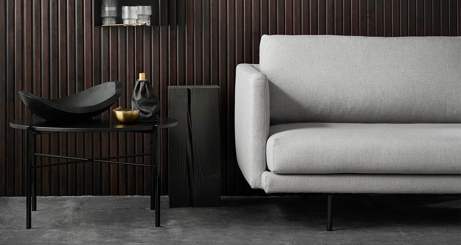 Harmaa sohva, musta metallijalkainen sivupöytä, tummat puupaneelit, musta ja kultainen asetelma