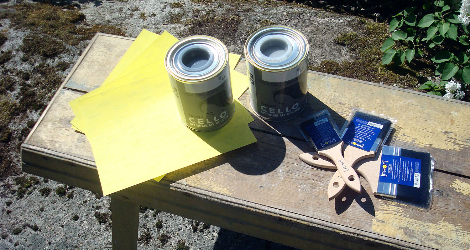 Puupöydällä ulkona kaksi avattua harmaamaalipurkkia, keltaiset paperiarkit ja puiset pensselit.