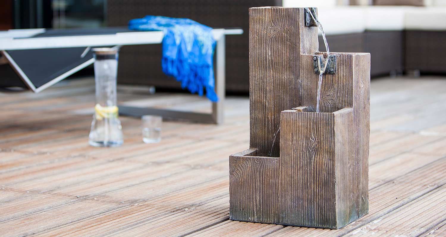 Puuaskelmista laskeva vesiaihe terassilla, taustalla sininen viltti ja sitruunavettä karahvissa.