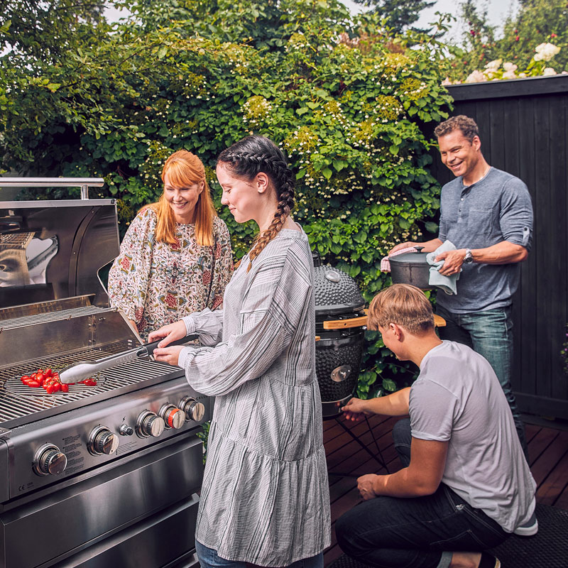 Neljä ihmistä puutarhassa grillaamassa tomaatteja kaasugrillissä, taustalla vihreää pensasta.