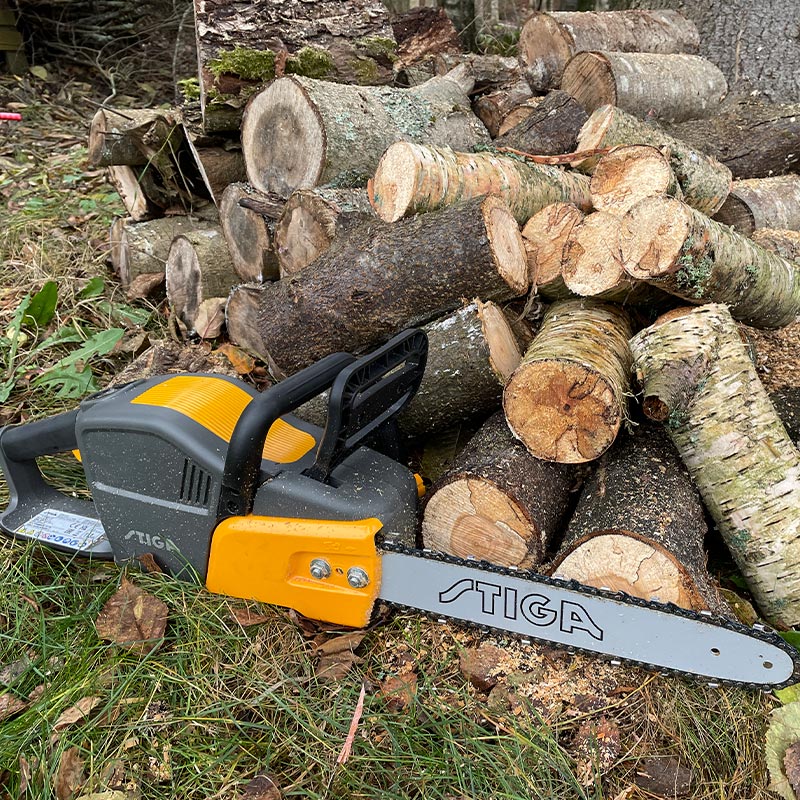 Stiga CS 700E -akkuketjusaha puupinon edessä ulkona, koivupuita ja muuta puuta sahattuna.