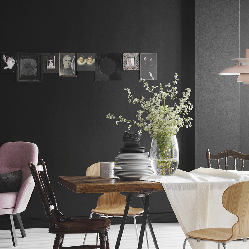 Ruokailutila tumma seinä, puupöytä valkoisella kaitaliinalla, maljakko valkoisin kukin, eri tuolit.