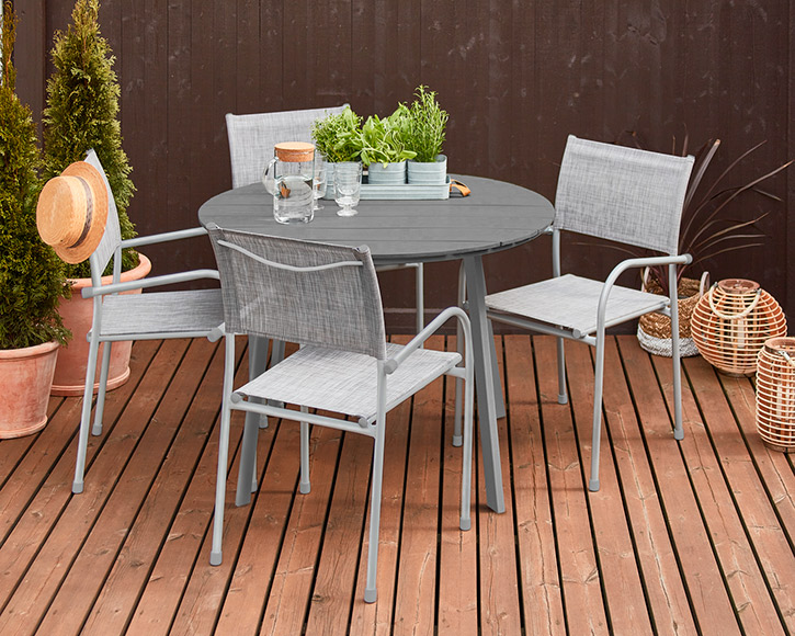 Pyöreä harmaa terassipöytä neljällä tuolilla puulattialla, yrttiruukku, vesikarahvi, olkihattu