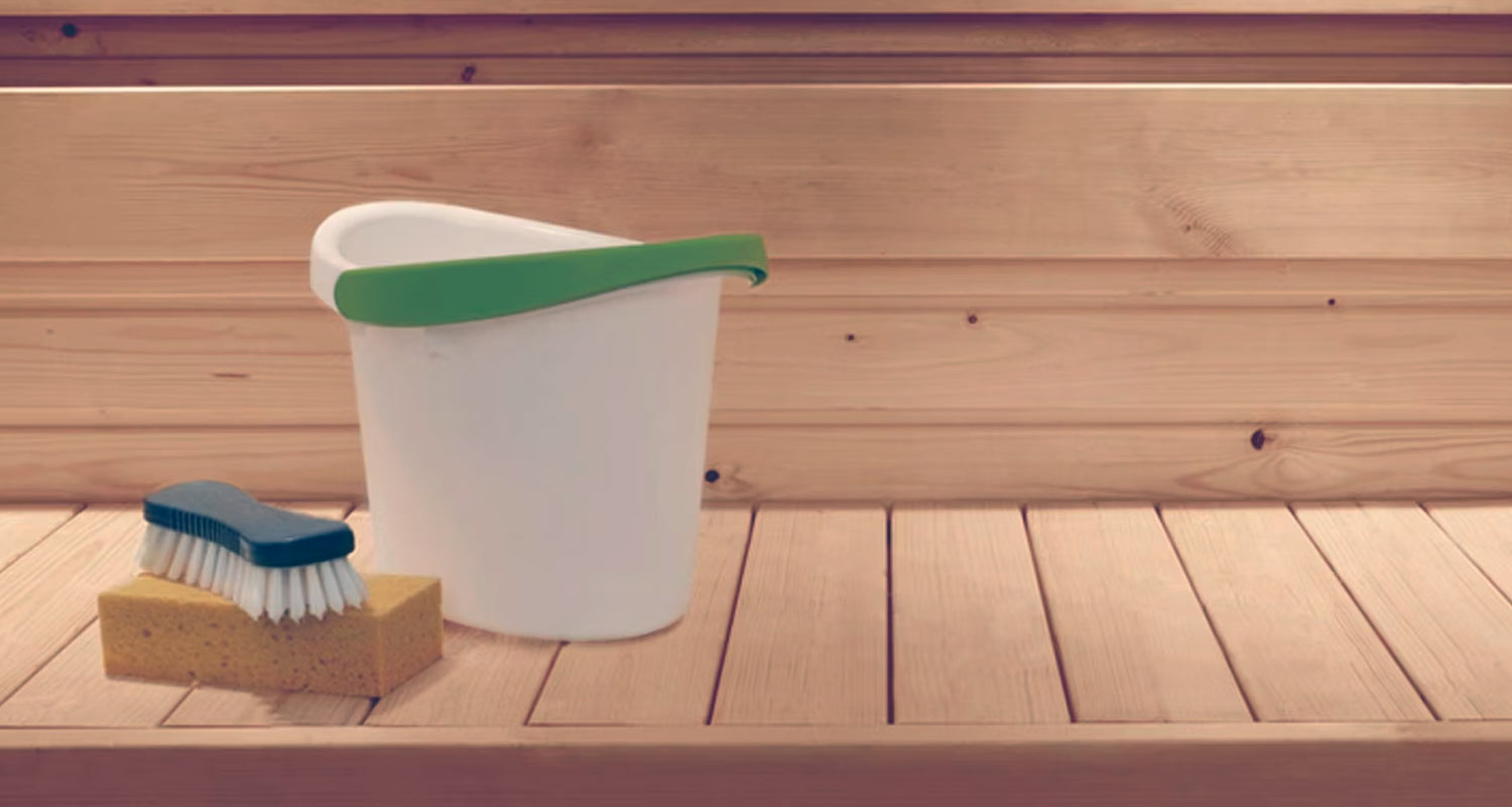 Valkoinen ämpäri vihreällä reunuksella, sininen harja keltaisen sienen päällä puupinnalla.