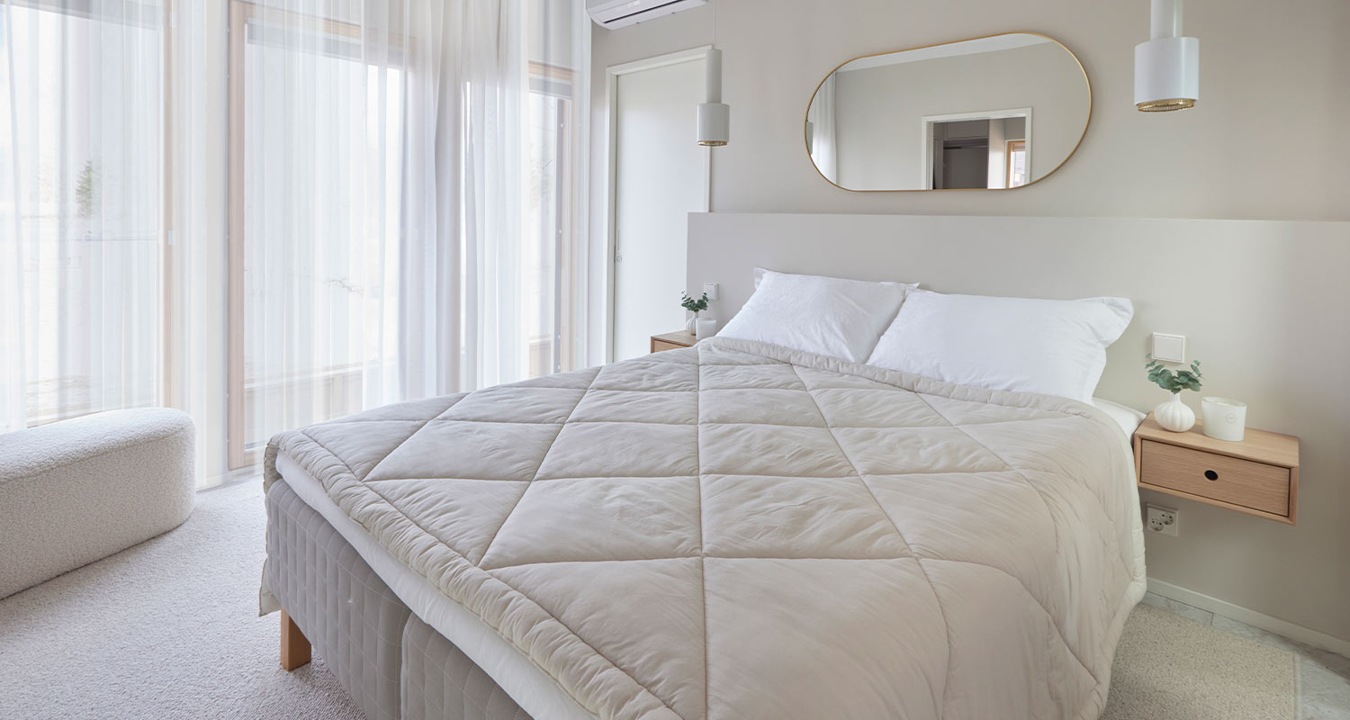 Vaalea skandityylinen makuuhuone, valkoinen leveä sänky beigen peiton ja kevyiden verhojen kera.