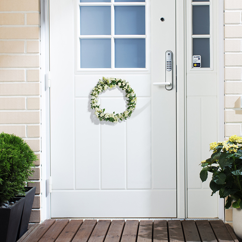 Valkoinen ulko-ovi sinisillä yläikkunoilla, kukkaseppele ja ruukkukasvit.