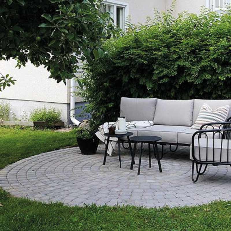 Harmaa kulmasohva ja mustat sivupöydät pyöreällä harmaalla laattapatiolla taustalla vihreää pensasta