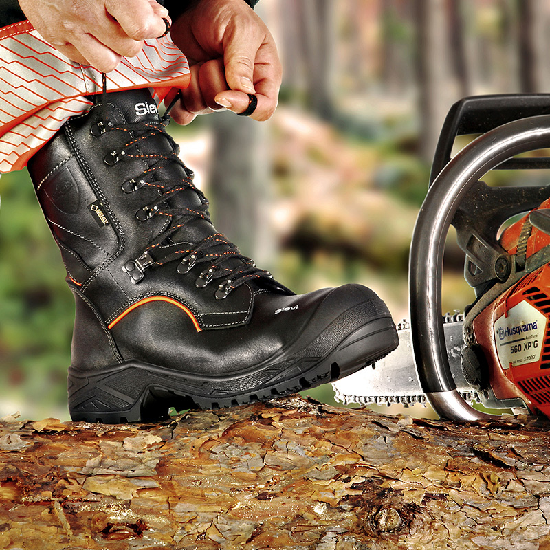 Musta oranssikoristeinen suojakenkä, käsi nyörejä solmimassa, moottorisaha ja puunrunko.