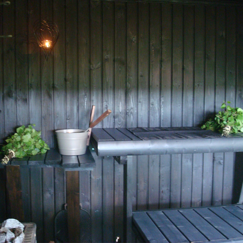 Tumma puusaunan lauteet, löylykiulu kauhalla ja vihdat lauteen reunalla.