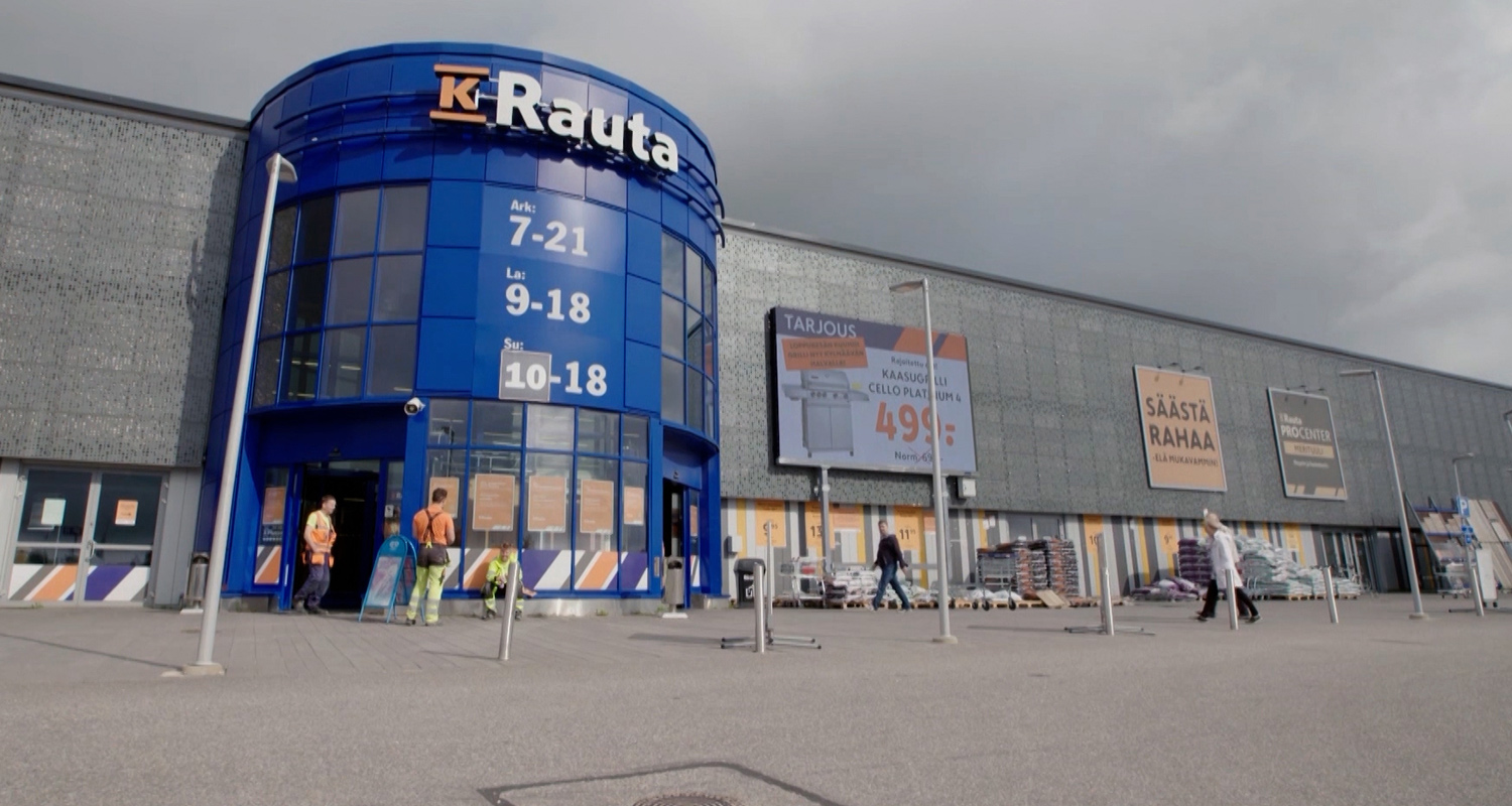 K-Raudan sininen sisäänkäyntitorniosa harmaan julkisivun edessä, pilvinen taivas ja työasuinen ryhmä.