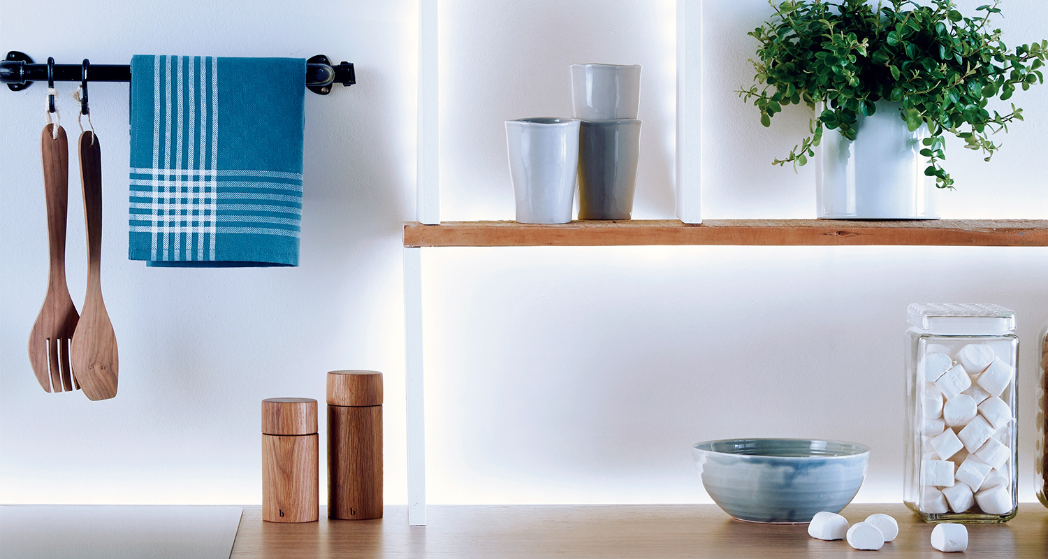Keittiöhylly: sinivalkoinen pyyhe, puiset työkalut, harmaat mukit, viherkasvi, kulho ja purkki