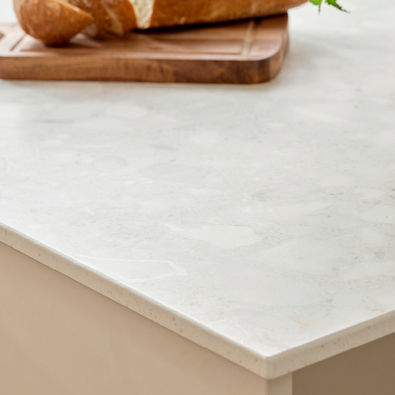 Vaalea marmorikuvioitu keittiötaso, reunalla puinen leikkuulauta, viipaloitua leipää ja yrtti.