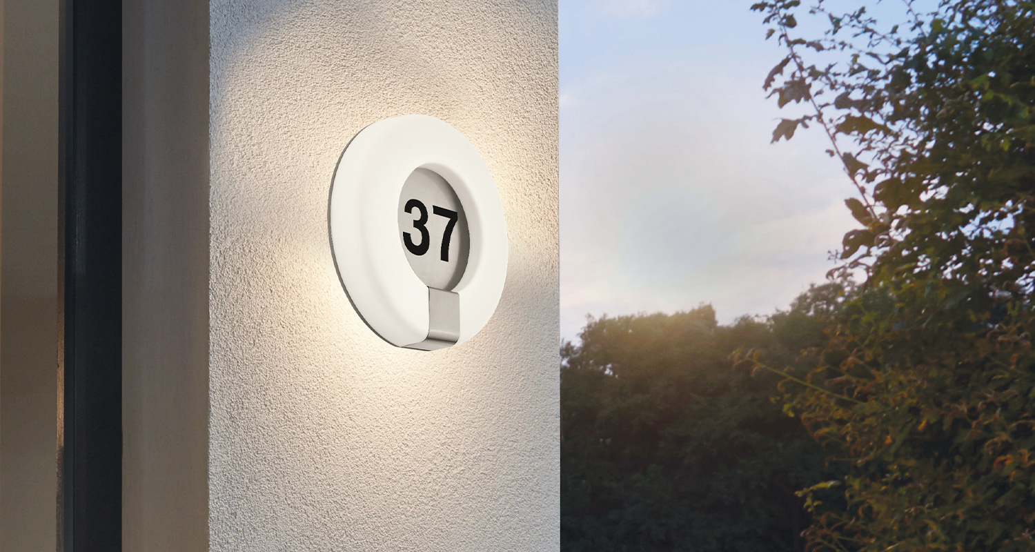 Pyöreä valkoinen lamppu mustalla numerolla 37, lämmin valo valkoisella seinällä ja hämärät puut.