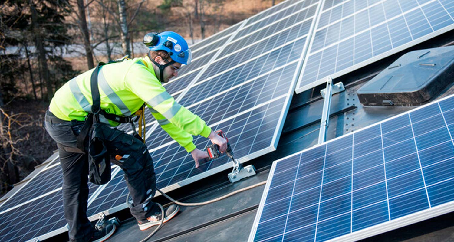 Katolla asentaja kiinnittää sinisiä aurinkopaneeleja neonvihreässä takissa sinisellä kypärällä