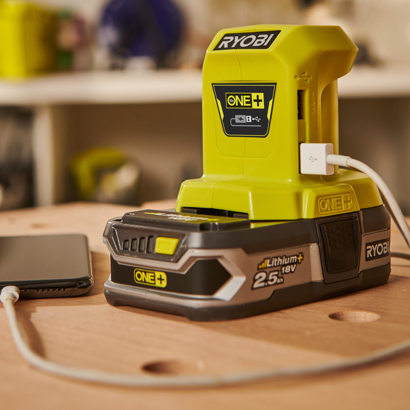 Neonvihreä Ryobi One+ 18V litiumakku USB-latausasemassa, puhelin liitettynä pöydällä
