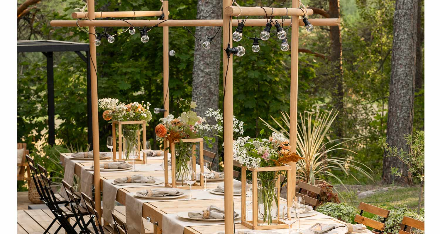 Ulkoterassilla pitkä vaalea puupöytä valkoisin liinoin, kukkia puisissa telineissä ja valosarjoissa.