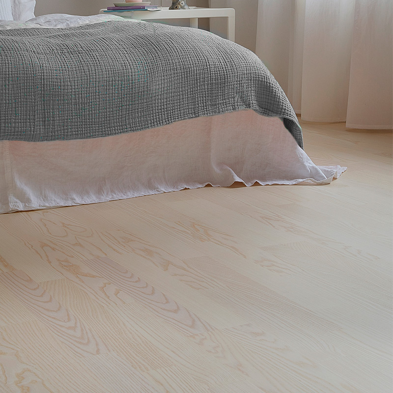 Valoisassa makuuhuoneessa vaalea puulattia, valkoinen lakanakangas ja harmaa täkki.