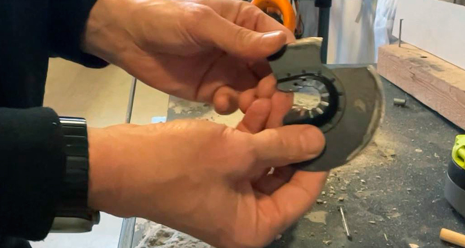 Kaksi kättä pitelee mustaa puolikuun muotoista teräsuojalevyä pölyisellä työpöydällä.