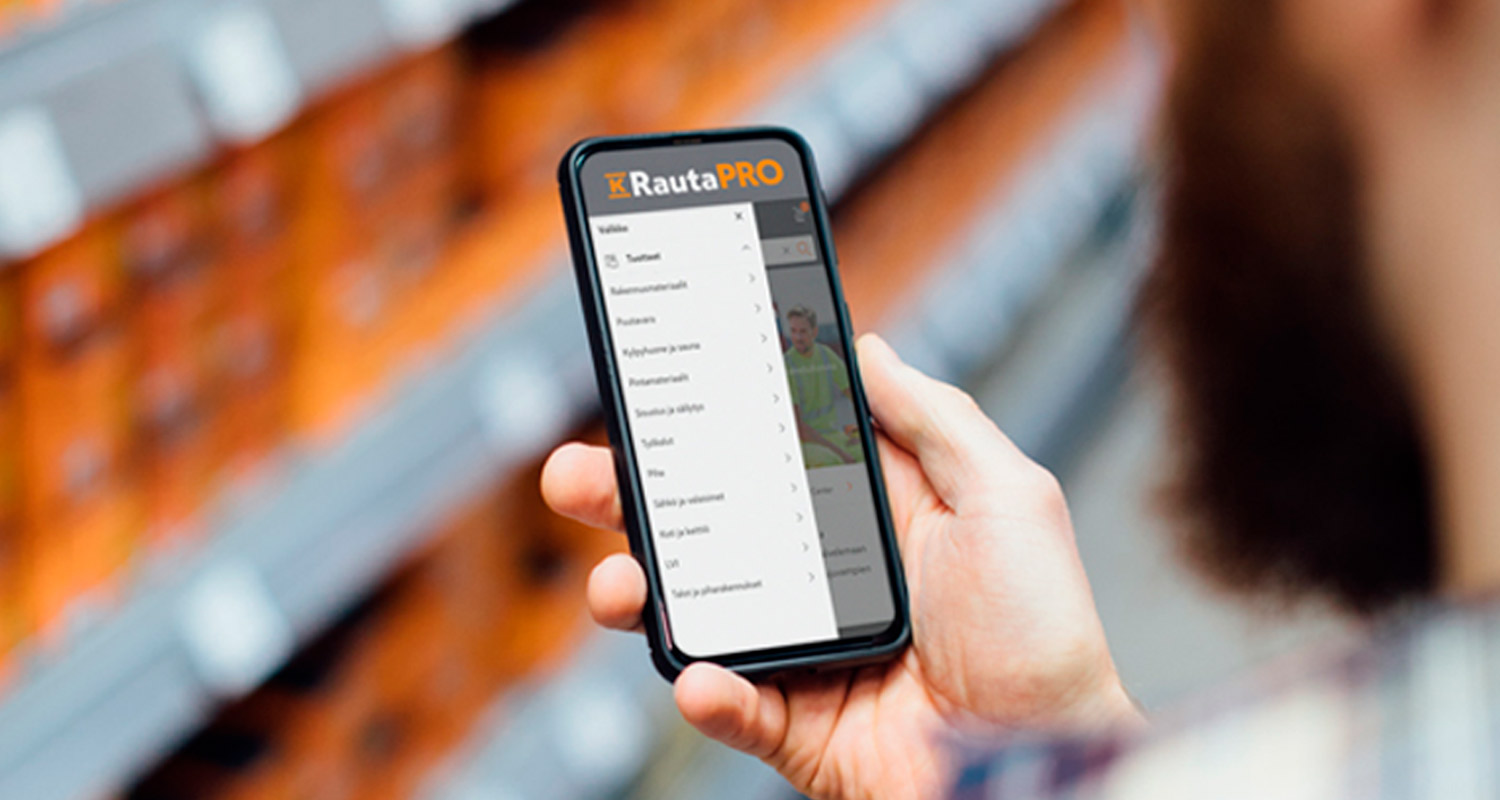 Käsi pitelee älypuhelinta rautakaupan oranssin hyllyrivin edessä, ruudulla RautaPRO-valikko.