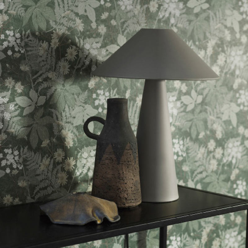Harmaa pöytälamppu, ruskea savivaasi ja metallilehti vihreää kukkakuvioista seinää vasten