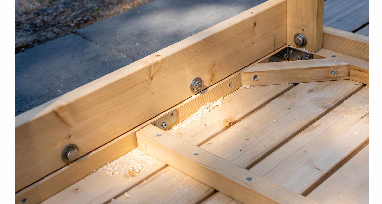 Puinen runkorakenne puulaudoista, metallikulmaraudat ja puulastuja auringonvalossa betonilla.