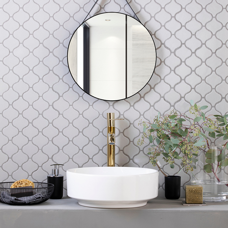 Pyöreä peili, valkoiset mosaiikkilaatat, messinkihana, valkoinen pesuallas ja vihreitä oksia.