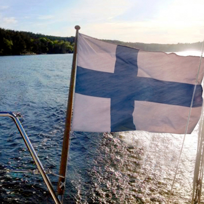 Veneen perässä liehuva sinivalkoinen Suomen lippu järvimaisemassa auringon kajossa