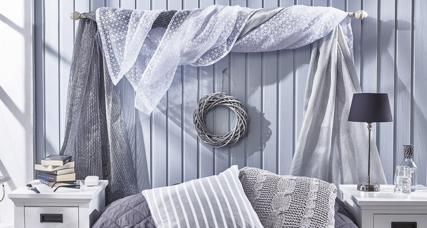 Sinivalkoisella seinällä harmaat ja valkoiset verhot, kranssi, tyynyt sängyllä ja yöpöytä lamppuineen.