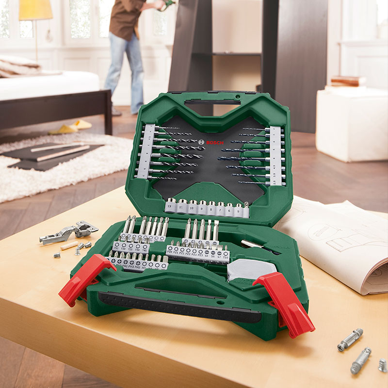 Vihreä Bosch-työkalulaukku auki pöydällä, poranteriä ja ruuvauskärkiä, taustalla huonekalun kasaaja.