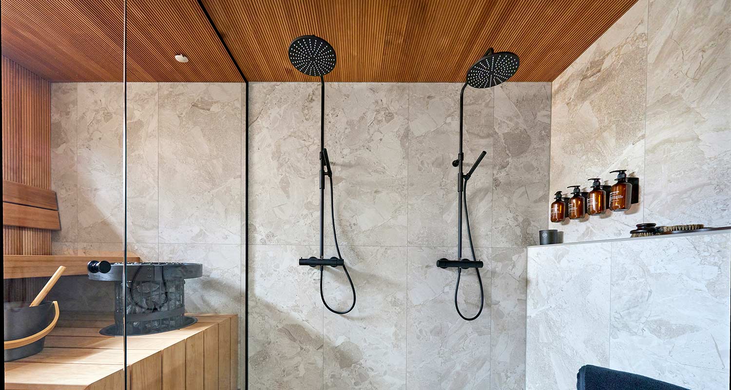 Saunallinen kylpyhuone: puuta, harmaata laattaa, 2 mustaa suihkua, ruskeat pullot