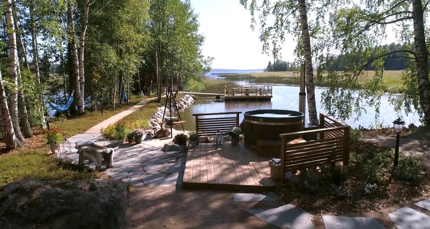 Koivikkoisen järvenrannan aurinkoinen terassi, ruskea puupalju ja penkit laiturilla.