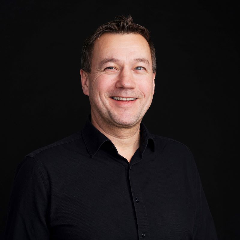 Rintakuva henkilöstä mustassa kauluspaidassa mustaa taustaa vasten.